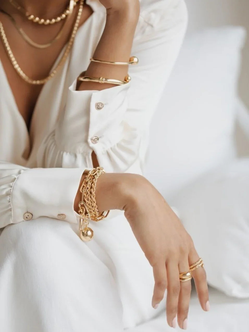 Mujer con ropa blanca y joyería dorada elegante: pulseras, anillos y collares en ambiente claro