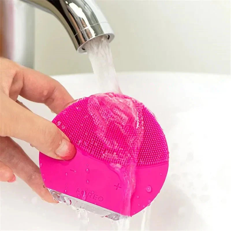 Mano sosteniendo cepillo facial eléctrico rosa Foreo bajo grifo con agua